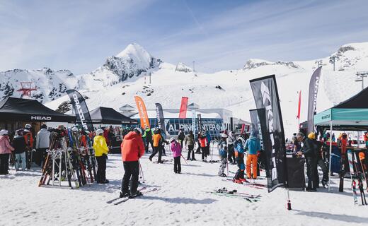 Skitest-Veranstaltung im Schnee vor Bergkulisse mit Menschen, Skiern und Werbeflaggen