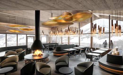 Interior view of a restaurant with fireplace, tables, chairs and gold-colored lamps under a wooden ceiling